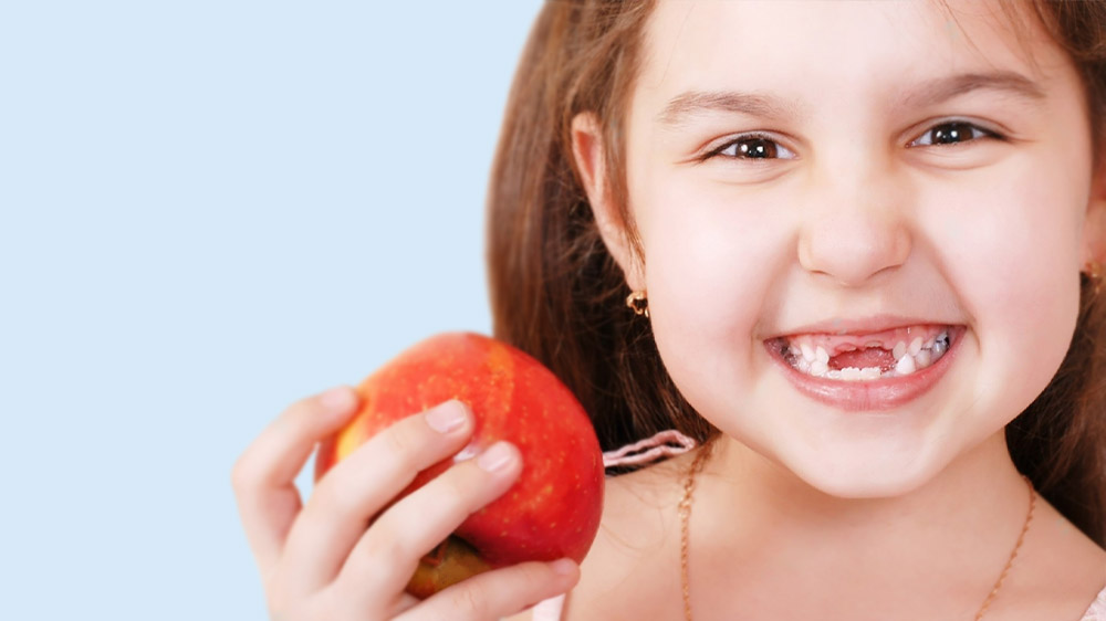 دختری که دندان های پیش را ندارد و یک سیب در دستش نگه داشته است.