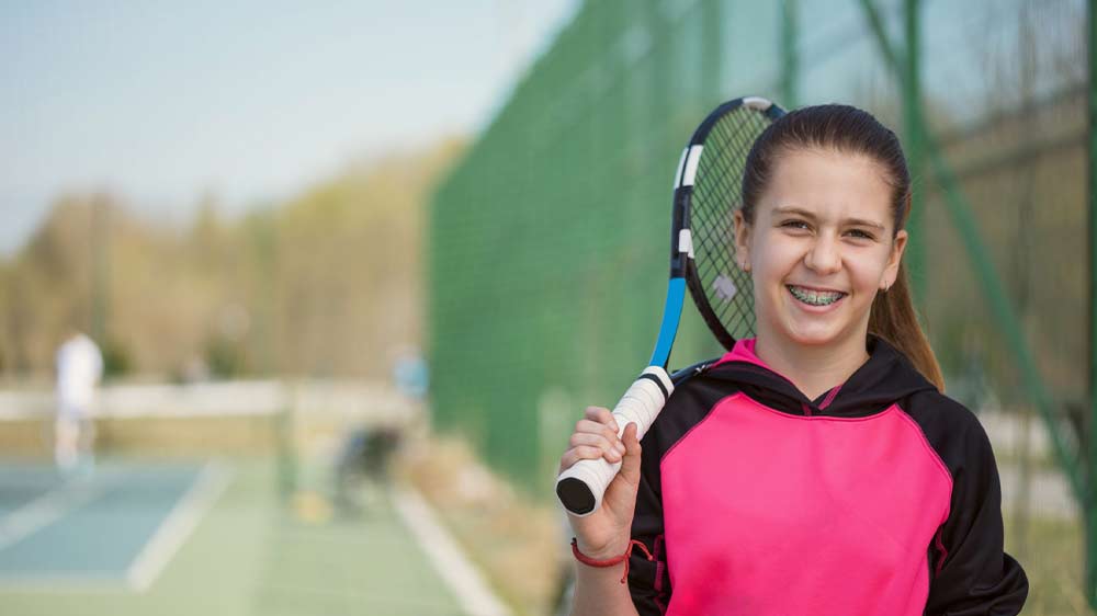 دختری با لبخند و دندان های ارتودنسی که راکت تنیس در دست دارد.
