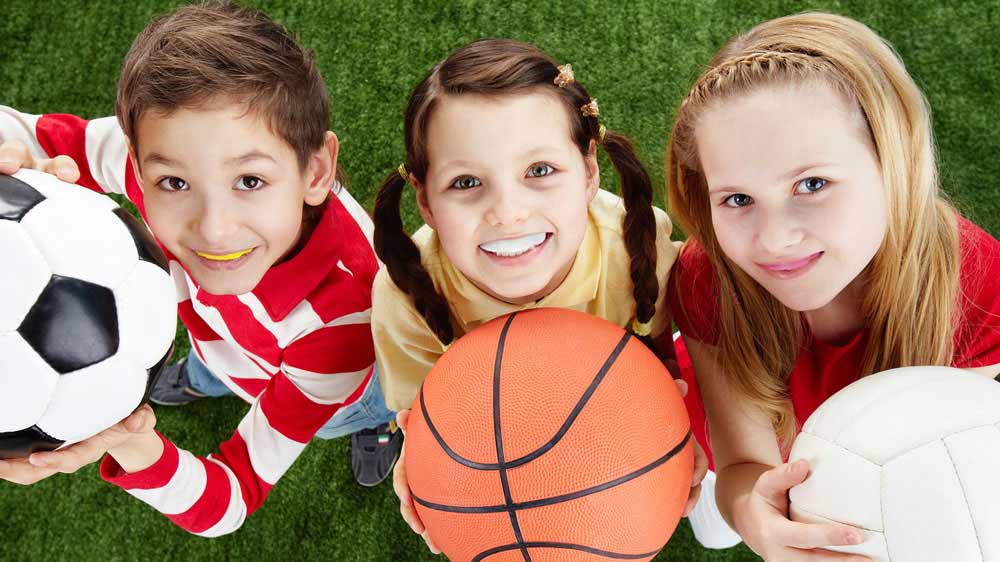 ثلاثة أطفال مبتسمون يحملون كرات رياضية ويرتدون واقيات الفم المخصصة لتقويم الأسنان أثناء ممارسة الرياضة.